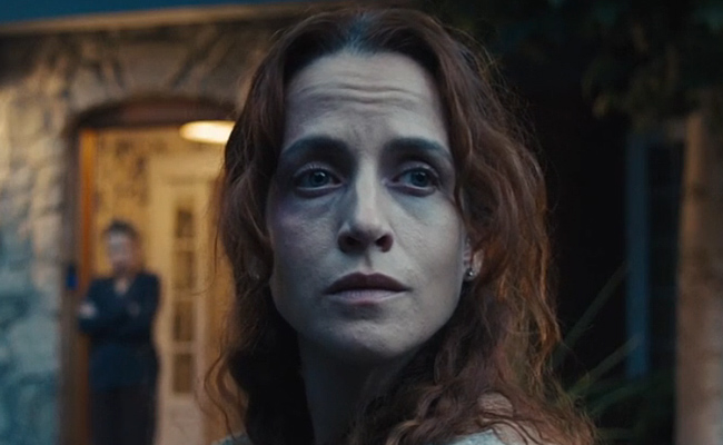 A close-up still of Kimberly-Sue Murray as Daisy with long, wavy reddish-brown hair looking off-camera with a concerned expression. In the softly blurred background, Diana Salvatore as Louise stands in the open doorway of a stone house at twilight.