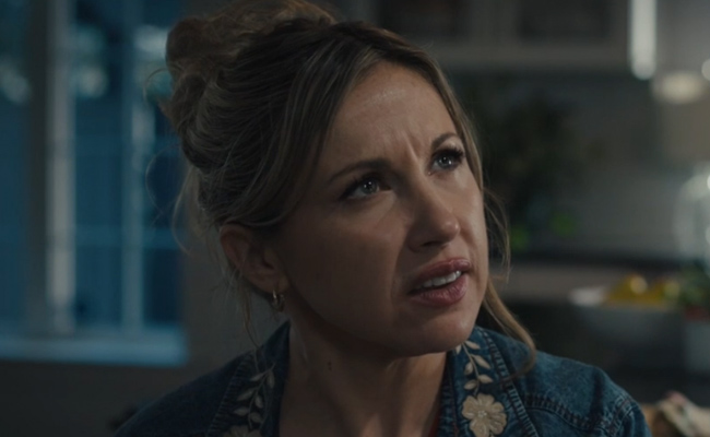 A close-up still of Diana Salvatore as Louise, a woman with blonde hair styled in a high bun, wearing an embroidered denim jacket, looking off-screen with a slightly concerned and questioning expression in a softly lit indoor setting.