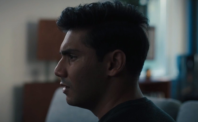 Side profile Ansh Pandey as Zack, a young man with dark hair looking off to the side with a pensive expression, captured in a dimly lit, softly blurred indoor environment.