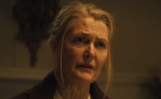 A close-up still of Mary Lewis as Mrs. Simpson, a woman with fair skin and light hair pulled back, wearing a dark shawl over a light-colored top. She has a concerned and slightly distressed expression on her face with visible wrinkles on her forehead, set against a dimly lit, out-of-focus interior background.