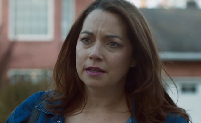 Close-up still of Jenny Pudavick as Morgan Marsh, a woman with shoulder-length brown hair and a concerned expression, wearing a denim jacket, set against a blurred residential background.
