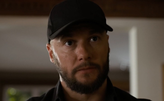 Close-up still of Dennis Andres as Grant Dreyer, a man with a short, trimmed beard wearing a black baseball cap and a black collared shirt, looking forward with a neutral, steady expression in an indoor setting.