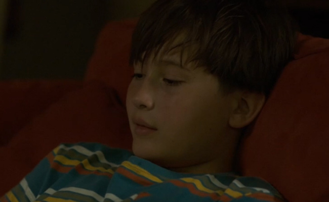 Jordan Mazeral as Cody Marsh, a young boy with short brown hair wears a teal shirt with horizontal yellow, white, and reddish-orange stripes while leaning against a dark red sofa in a dimly lit room.
