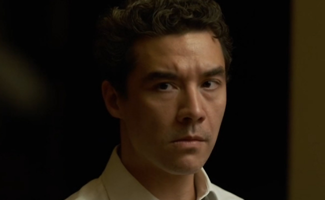A medium close-up stil of Chris Hong as Kevin Marsh, a man with dark wavy hair and a serious, focused expression, wearing a white collared shirt against a dark, dimly lit background.