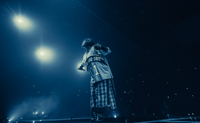 From the 2026 Billie Eilish concert film, "Hit Me Hard and Soft: The Tour Live in 3D", Eilish wears a sports jersey, with "Hard and Soft 01" on the front, and plaid pants on a dimly lit stage, illuminated by bright spotlights and a scattering of smaller lights in the background.