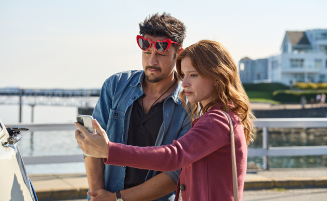 Tyler Hynes as Mark. a man wearing red heart-shaped sunglasses and a blue shirt stands beside Stacey Farber as Amy, a woman with wavy reddish-brown hair wearing a pink sweater, both looking closely at a smartphone she is holding in an outdoor, sunny coastal setting with a pier visible in the background.