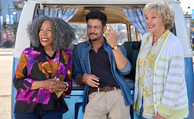 Three people stand in front of an open, light blue and white vintage camper van. BJ Harrison as Sue, a black woman with curly gray hair and a vibrant patterned jacket stands on the left. In the center, Tyler Hynes as Mark, a man with a beard, wearing a denim shirt and beige trousers, gestures while speaking to the woman on the right, Christine Ebersole as Vivien, who has short blonde hair and wears a white cardigan over a yellow patterned top.