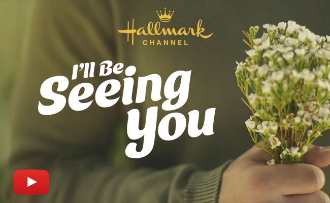A person wearing a green sweater holds a small bouquet of white wildflowers against a soft, out-of-focus background. The Hallmark Channel logo is positioned at the top, while the title "I'll Be Seeing You" appears in a large, white script font across the center. A red YouTube play button icon is visible in the bottom left corner.
