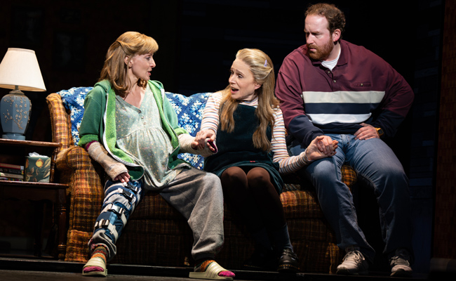 Three individuals are seated on a couch, holding hands, in what appears to be a theatrical production or scene. The person on the left, Laura Woyasz as Pattie Levaco, visibly pregnant and wearing a green hooded sweatshirt over a patterned dress, has a brace on right leg, and casts on both arms. The person in the middle, Ann Morrison as Kim Levaco, wearing a green jumper dress over a striped shirt, also appears to be interacting emotionally. The person on the right, Jim Hogan as Buddy Levaco, wearing a striped polo shirt and jeans, looks towards his right. A lamp and a side table with books and a tissue box are visible to the left side of the image.