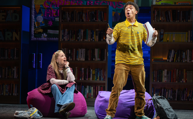 A young man in a yellow striped shirt, Marcus Phillips as Seth Weetis, gestures expressively while holding a paper, standing next to a woman, Ann Morrison as Kim Levaco, in a burgundy top and jeans, seated on a large beanbag chair, with bookshelves in the background of a stage set of a school library.