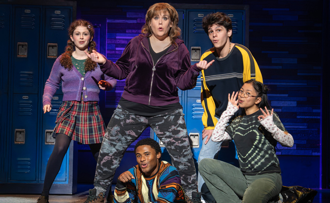 Five performers in casual, colorful attire pose in front of blue lockers, with one in the foreground and four behind. From left in the rear, Skye Alyssa Friedman as Teresa, wearing a purple cardiagn sweater over a green top, with a plaid skirt and black leggings, her reddish hair in two pigtails. In the center rear is Emily Kock as Debra, in a purple velour zipped jacket, black V-neck shirt, and camouflage leggings. Rear right is Max Santopietro as Aaron, in a yellow and black oversized sweater and blue denim jeans. Bottom center is Darron Hayes as Martin, in a multi-colored sweater. Bottom right is Gabby Beredo as Delia, wearing a faded black T-shirt with tie-dyed off-white hearts, a white patterned long-sleeved shirt and denim blue jeans.