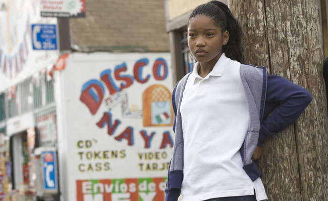 From "Akeelah and the Bee", a young Black girl (Keke Palmer as Akeelah) with braided hair pulled back in a ponytail stands leaning against a wooden post. She wears a white polo shirt and a blue and grey zip-up hoodie draped over her shoulders. In the background, there is a brightly painted mural on the side of a building with images of a jukebox and a flag. A blue sign with a white telephone symbol is also present.