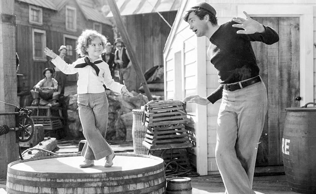 From "Captain January", a black-and-white image of a young girl (Shirley Temple) in sailor attire dances on a barrel while a man (Buddy Ebsen) in a sailor outfit dances along, with a nautical-themed set in the background featuring lobster traps and buildings.