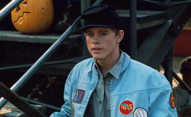 From "Eat My Dust!", a young man (Ron Howard) wearing a black cap and a light blue denim jacket with patches stands next to a staircase with a carved pumpkin on a step.