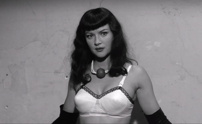 From "The Notorious Bettie Page", a black-and-white image of a woman (Grethcen Mol as Bettie Page) wearing a white brassiere, black opera gloves, and a ball gag around her neck like a necklace. She has dark, wavy hair with bangs and is looking directly at the viewer. The background is a textured, plain wall.