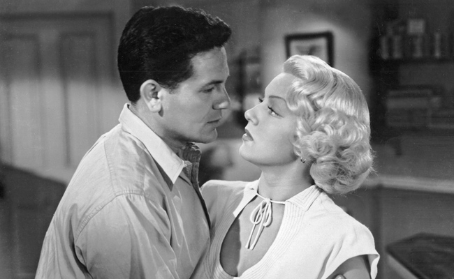 From "The Postman Always Rings Twice", a black-and-white image of a man and a woman gazing intensely at each other in an intimate moment. The man ( (John Garfield as Frank Chambers) has dark, slicked-back hair and wears a light-colored collared shirt. The woman (Lana Turner as Cora Smith) has blonde, curled hair and wears a light-colored top with a V-neckline and a delicate tie around her neck. The background is softly blurred, suggesting an indoor setting with hints of a framed picture and shelving.