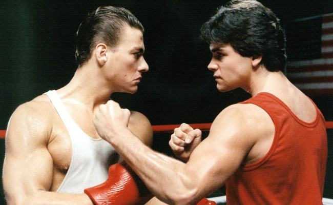 From "No Retreat, No Surrender", two muscular young men in boxing gear face each other in a boxing ring, their fists raised in anticipation. The man on the left (Jean-Claude Van Damme as Ivan Kraschinsky the Russian) has dark, slicked back hair and wears a white tank top with red boxing gloves. The man on the right (Kurt McKinney as Jason Stillwell) has dark hair and wears a red tank top, but is bare-knuckled. The men are in a boxing ring, an American flag visible on the right in the dark background.