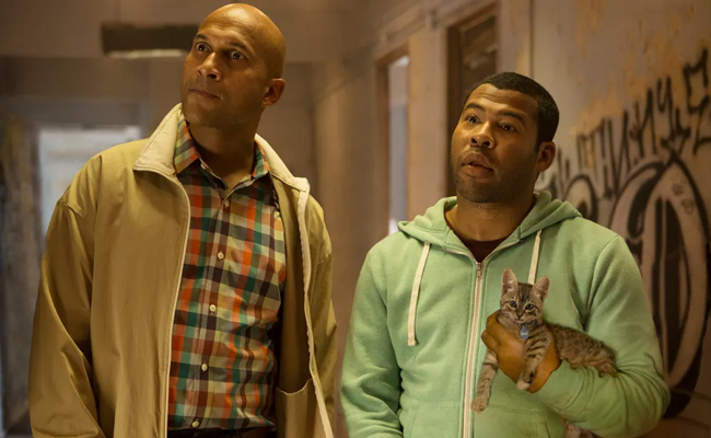 From "Keanu", two men stand in a dimly lit hallway with graffiti on the wall, one holding a tabby kitten. The man on the left (Keegan-Michael Key as Clarence Goobril) is bald, wears a tan jacket and plaid shirt. The man on the right holding the kitten (Jordan Peele as Rell Williams), has dark, short hair, a lime green hoodie over a black T-shirt. Both have confused looks on their faces.