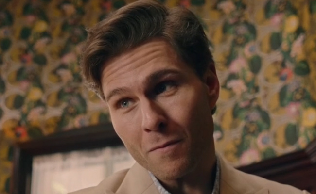 John Ryan McLaughlin as Alec. Close-up of a man with neatly styled brown hair, wearing a light beige blazer over a patterned shirt, set against a background of vintage floral wallpaper with yellow and green accents.