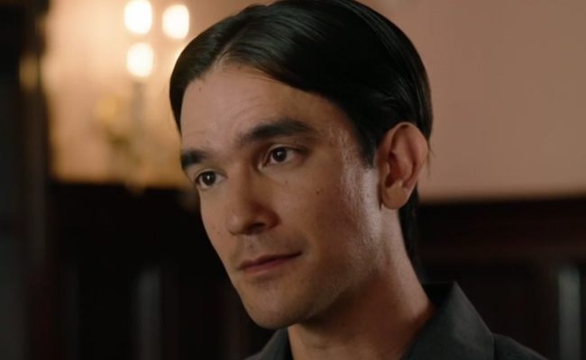 James Chrosniak as Jasper. A close-up portrait of a man with dark hair parted in the center, wearing a dark collared shirt. He is looking off-camera with a neutral, contemplative expression against a softly lit, out-of-focus indoor background.