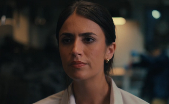 A woman in a kitcken with dark hair pulled back, wearing a white shirt, looks intently forward with a neutral expression.