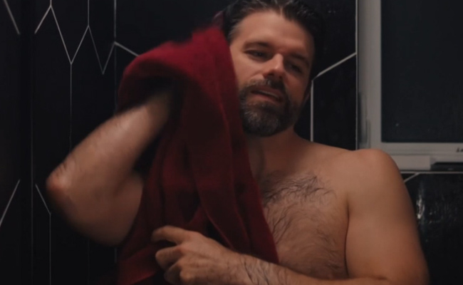 A man with a beard uses a large red towel to dry his hair while standing in a shower with dark geometrically patterned wall tiles.
