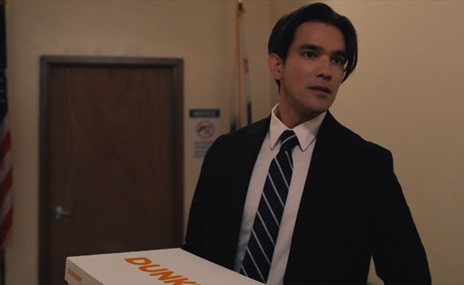 A man with dark hair, wearing a black suit and a blue and white striped tie, holds a white Dunkin' box while looking to the side in an indoor office setting.