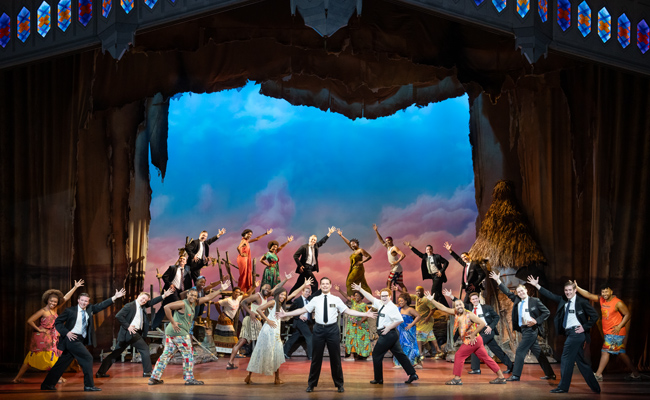 A large cast of performers stand on a stage with a backdrop of a blue sky, pink clouds, and a thatched hut. The performers are posed with arms outstretched, featuring a mix of actors in formal black suits with white shirts and ties, and others in colorful, patterned casual attire. A man in a crisp white short-sleeved shirt and black tie stands centered in the foreground as the focal point.