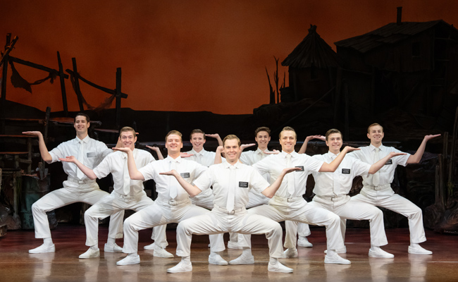 A group of nine men in identical white short-sleeved shirts, white ties, and white trousers perform a synchronized pose on a theatrical stage. Each man wears a small black nametag on his left chest and holds his arms outstretched with palms facing upward. They are positioned against a backdrop depicting a desolate landscape with rustic huts under a vibrant orange sky.