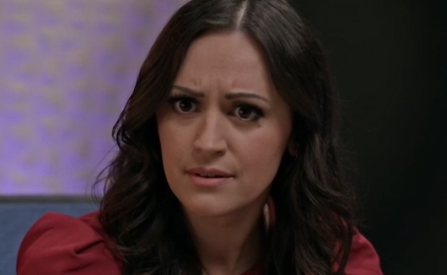 Harley Bronwyn as May. A person with long, wavy dark hair and a concerned, confused expression, wearing a red shirt, shown in a close-up shot against a dark, purple-lit background.