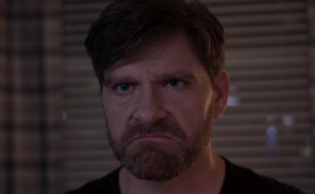 Clayton Louis as Zeke. A close-up still of a man with dark hair and a short beard, wearing a black shirt, looking toward the camera with a stern, furrowed expression in a dimly lit room with window blinds and plaid curtains visible in the background.
