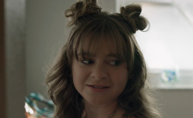 Lauren Cole as Poppy. A person with wavy, shoulder-length hair styled into two space buns on top of their head with soft bangs. She is looking off to the side with a skeptical, slightly unsure facial expression.