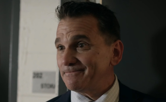 John Hardy as Detective Doye. A close-up still of a middle-aged man with dark hair wearing a navy suit and a light-colored checkered dress shirt, looking towards the side with a mischievous smirk and raised eyebrows. The background features a white wall with a small sign and a dark door frame.