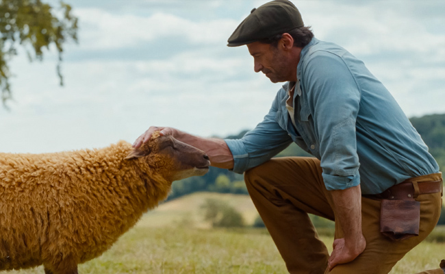 Hugh Jackman as George Hardy, wearing a flat cap and blue shirt gently stroking the head of a tan sheep in a grassy field with rolling hills in the background.