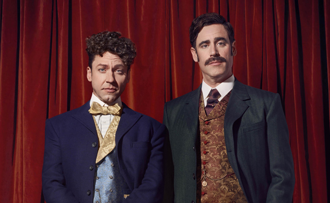 Promotional still image from "Houdini & Doyle". Two men in ornate, formal wear stand against a dark red velvet curtain backdrop. The man on the left, Michael Weston as Harry Houdini, wears a dark blue suit jacket with a light blue and gold patterned vest, a white shirt, and a large gold bow tie. He has curly dark hair and blue eyes. The man on the right, Stephen Mangan as Arthur Conan Doyle, wears a dark green suit jacket over a patterned brown and gold brocade vest, a white shirt, and a dark tie. He has dark hair styled back and a prominent mustache. Both men have serious expressions.