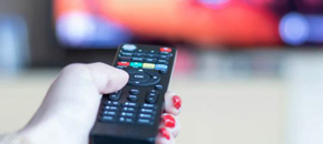 TV by the Decade featured image. A hand with red-painted fingernails holds a black television remote control aimed toward a blurred television screen in the background.