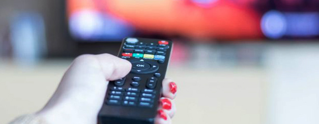 TV by the Decade featured image. A hand with red-painted fingernails holds a black television remote control aimed toward a blurred television screen in the background.