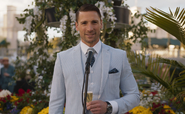 From "Kentucky Roses" Andrew Walker as Ash Taylor, a man in a blue and white pinstripe suit holding a champagne flute, speaking into a microphone, with floral decorations and palm fronds in the background.