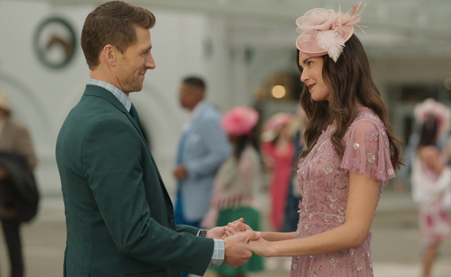 From "Kentucky Roses", Andrew Walker as Ash Taylor on the left, wearing a green suit, holds hands with Odette Annable as Sadie Green, wearing a pink dress and fascinator, blurred figures in the background at Churchill Downs on the day of the Kentucky Derby.
