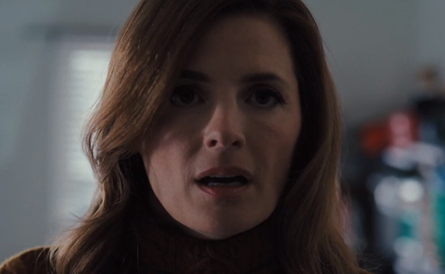 Close-up portrait of Stana Katic as Martha Carelli, a woman with wavy brown hair wearing a textured brown sweater, looking forward with a focused, slightly surprised expression and her mouth slightly parted.