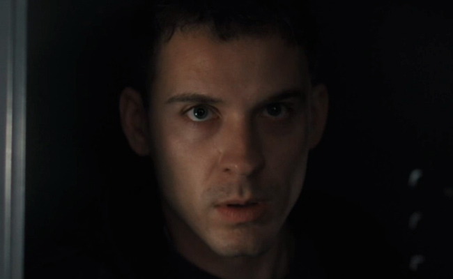 A close-up shot of Tyler Tom&aacute;s Perez as Michael Anderson, a man with short dark hair and light eyes looking directly at the camera with an intense, focused expression. The background is completely dark, and the lighting is dim and dramatic, highlighting the details of his face.