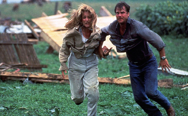 From "Twister", a man and a woman running frantically through a field of debris from a tornado, with the woman (Helen Hunt as Dr. Jo Harding) holding onto the man's (Bill Paxton as Bill Harding) arm. Hunt wears a beige, buttoned shirt over a white tank top, and faded denim jeans. Paxton wears a blue denim, buttoned shirt over a blue T-shirt, with dark blue denim jeans.