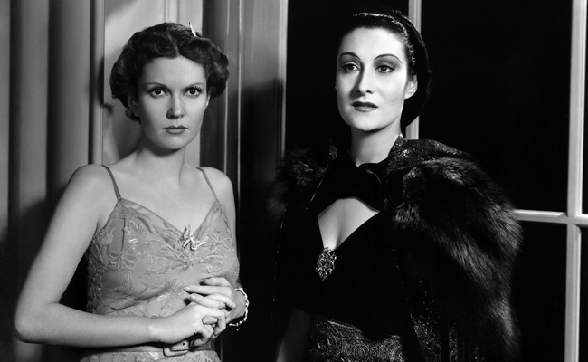 From "Dracula's Daughter", black-and-white image of two women in glamorous vintage attire standing by a window. The woman on the left (Marguerite Churchill as Janet Blake) wears a light-colored, embroidered slip dress with a butterfly brooch, the other (Gloria Holden as Countess Marya Zaleska) wears a dark velvet dress with a fur stole.
