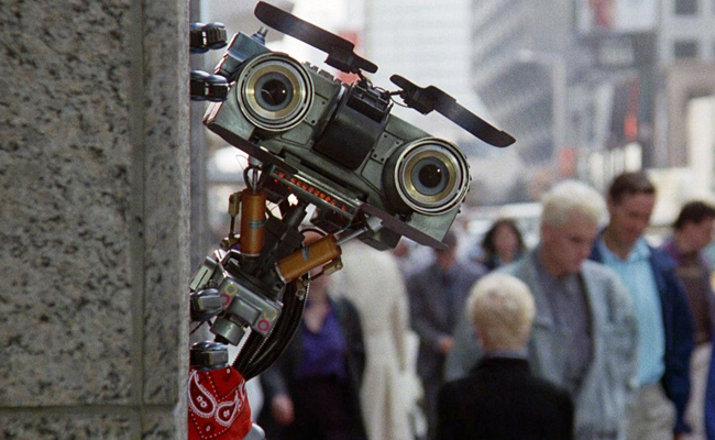 From "Short Circuit", a small, boxy robot with camera lenses for eyes, and a red bandana tied around its neck, peeks around a stone pillar into a blurred urban street scene with a crowd of people in the background.