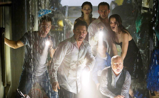 From "Poseidon", a group of people (Mike Vogel, Kurt Russell, Emmy Rossum, Josh Lucas, Jimmy Bennett, Jacinda Barrett, Richard Dreyfus) huddle together in a dark, wet environment, illuminating their faces with flashlights as water cascades around them. Vogel wears a grey T-shirt and denim jeans, Russell is in a white tux shirt and black pants, Lucas is wearing a formal white shirt, Rossum's gown is obstructed, Barrett wears a black, strapless gown, Dreyfus wears a gray formal shirt and black vest, Bennett, wears a blue buttoned shirt over a white T-shirt and black pants.