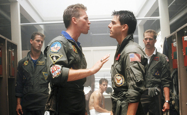 Still from "Top Gun" features Val Kilmer as Iceman and Tom Cruise as Maverick, dressed in flight suits and facing each other in a locker room during an argument. To the left is Rick Rossovich as Slider, and Anthony Edwards as Goose, both in flight suits, watching the argument. In the middle is Adrian Pasdar as Chipper, clad only in a white towel and sitting on a bench, also observing the argument.