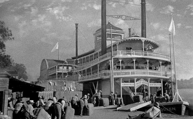 Still from "Show Boat" features a black-and-white image of a multi-decked paddlewheel steamboat docked at a riverbank. Large bales of cotton are piled high on the dock and being loaded onto the boat by a group of men. The steamboat, labeled "LOUISVILLE RIVER QUEEN NEW ORLEANS", has ornate railings and two tall smokestacks. The sky is overcast with dramatic clouds.