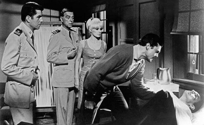 Still from "The Navy vs. the Night Monsters" features a black-and-white image of four people in a dimly lit room; two men in military-style uniforms, Anthony Eisley as Lt. Charles Brown and Phillip Terry as Base Doctor, stand to the left, looking at a woman, Mamie Van Doren as Nora Hall, with blonde hair in a sleeveless top in the center, while another man, Edward Faulkner as Bob Spaulding, in a V-neck sweater and collared shirt, bends over a person lying on a bed to the right.