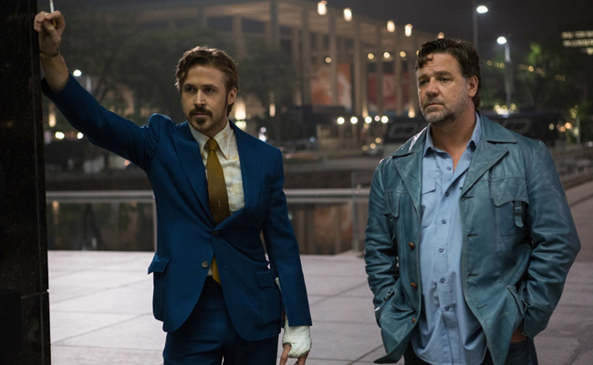 Still from "The Nice Guys" features Ryan Gosling with a mustache as Holland March standing to the left, in a blue two-piece suit with white shirt and gold necktie, his right arm up, holding a cigarette and leaning against the corner of a wall, his left hand in a white cast, and Russell Crowe with stubble as Jackson Healy wearing a light blue leather jacket, a light blue Oxford shirt and blue denim jeans. A blurry city is in the background.