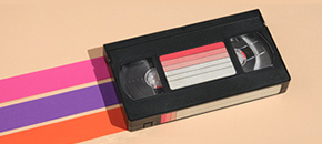 A black VHS tape placed diagonally on a light tan surface, featuring a label with horizontal red and white gradient stripes. Behind the tape are three parallel, vibrant bands of pink, purple, and orange extending across the frame.